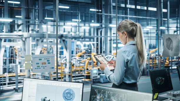 istock Car Factory Office Concept: Female Chief Automotive Engineer Using Tablet Computer to Develop, Design and Optimize Efficiency of Automated Robot Arm Assembly Line Manufacturing Electric Vehicles. 1352825040
