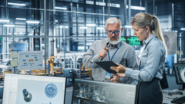 istock Car Factory Office: Scientist Talks with Senior Engineer, Use Tablet Computer to Design Production Conveyor for Advanced Power Engines. Automated Robot Arm Assembly Line Manufacturing Vehicles 1352825046