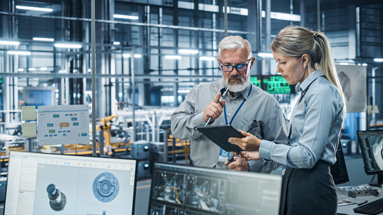 Car Factory Office: Scientist Talks with Senior Engineer, Use Tablet Computer to Design Production Conveyor for Advanced Power Engines. Automated Robot Arm Assembly Line Manufacturing Vehicles