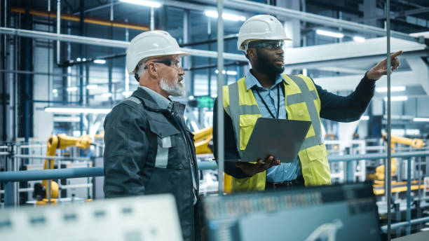 istock Car Factory: Project Manager and Automotive Engineer in Hard Hat, Talking, Using Laptop. Monitoring, Control, Equipment Production. Automated Robot Arm Assembly Line Manufacturing Electric Vehicles 1352825054