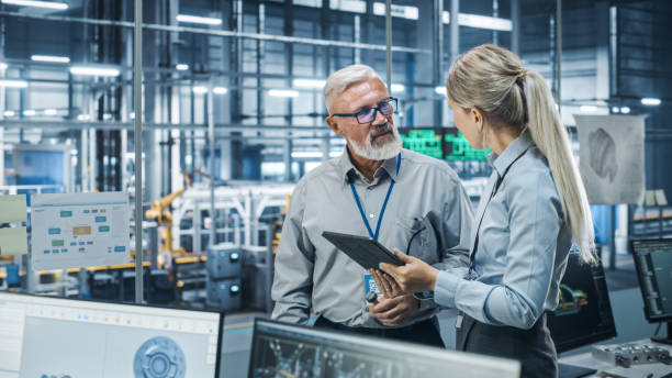 istock Car Factory Office: Scientist Talks with Senior Engineer, Use Tablet Computer to Design Production Conveyor for Advanced Power Engines. Automated Robot Arm Assembly Line Manufacturing Vehicles 1352825056