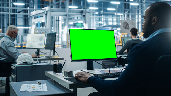 Car Factory Office: Black Male Chief Automotive Engineer Sitting at His Desk Working on Green Screen Chroma Key Computer. Automated Robot Arm Assembly Line Manufacturing. Over Shoulder Shot
