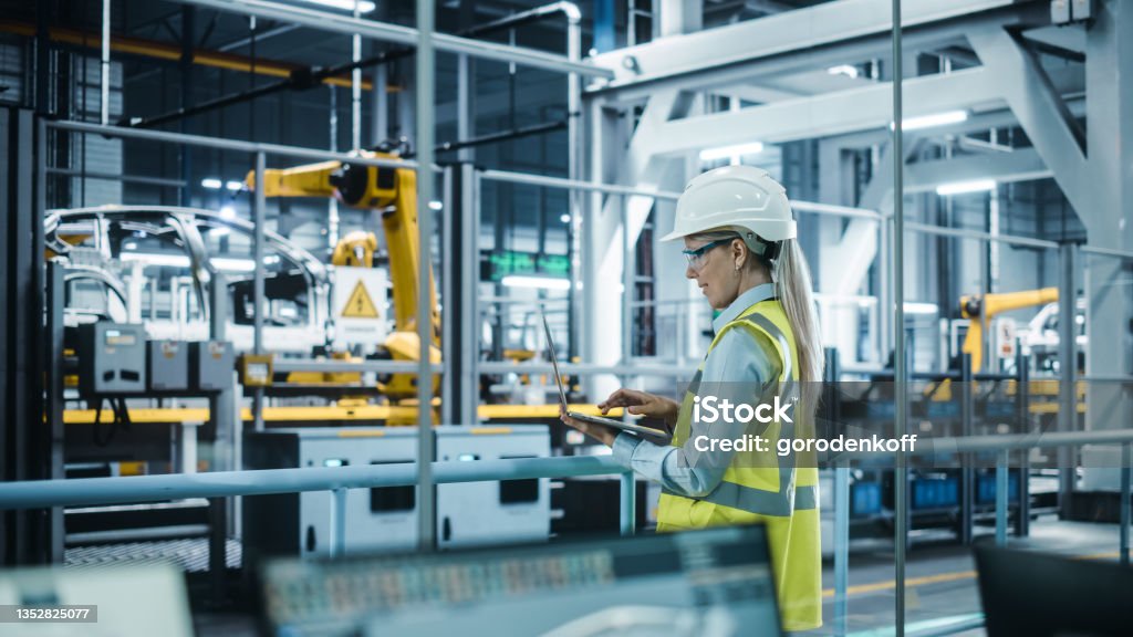 Car Factory: Female Automotive Engineer Wearing Hard Hat, Standing, Using Laptop. Monitoring, Control, Equipment Production. Automated Robot Arm Assembly Line Manufacturing Electric Vehicles. Manufacturing Stock Photo