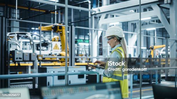 Car Factory Female Automotive Engineer Wearing Hard Hat Standing Using Laptop Monitoring Control Equipment Production Automated Robot Arm Assembly Line Manufacturing Electric Vehicles Stock Photo - Download Image Now