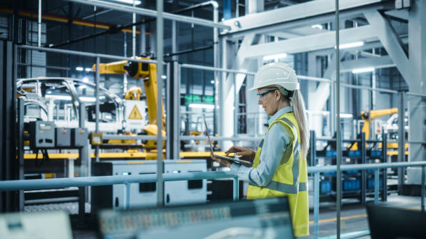 car factory: female automotive engineer wearing hard hat, standing, using laptop. monitoring, control, equipment production. automated robot arm assembly line manufacturing electric vehicles. - indústria imagens e fotografias de stock