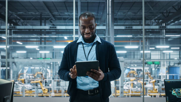 istock Car Factory Office: Portrait of Successful Black Male Chief Engineer Using Tablet Computer in Automated Robot Arm Assembly Line Manufacturing High-Tech Electric Vehicles. Medium Shot 1352825124