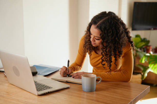 istock Multi-cultural female focussed, writing in notebook. Young professional remote working with laptop in modern apartment 1352825135