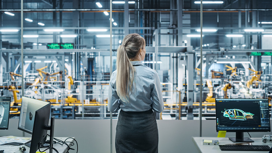 Car Factory Office: Successful Female Chief Engineer Overlooking Factory Production Conveyor. Automated Robot Arm Assembly Line Manufacturing Advanced High-Tech Electric Vehicles. Back View Shot