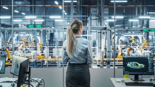 Photo of Car Factory Office: Successful Female Chief Engineer Overlooking Factory Production Conveyor. Automated Robot Arm Assembly Line Manufacturing Advanced High-Tech Electric Vehicles. Back View Shot