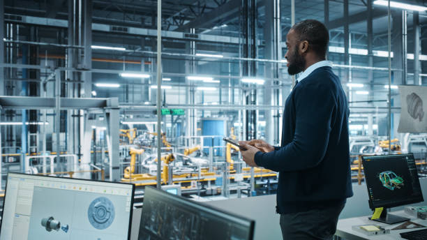 istock Car Factory Office: Portrait of Successful Black Male Chief Engineer Using Tablet Computer in Automated Robot Arm Assembly Line Manufacturing High-Tech Electric Vehicles. Side View Shot 1352825149