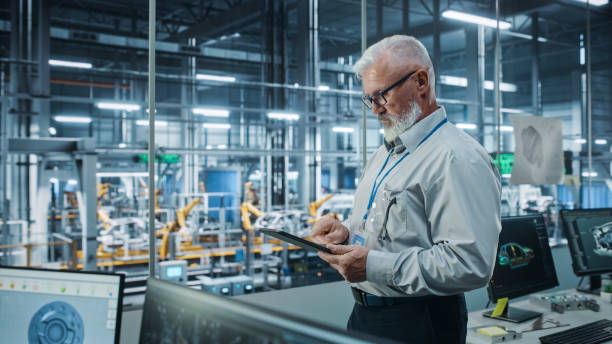 Car Factory Office: Portrait of Senior White Male Chief Engineer Using Tablet Computer in Automated Robot Arm Assembly Line Manufacturing High-Tech Electric Vehicles. Side View Shot Car Factory Office: Portrait of Senior White Male Chief Engineer Using Tablet Computer in Automated Robot Arm Assembly Line Manufacturing High-Tech Electric Vehicles. Side View Shot process automation office stock pictures, royalty-free photos & images