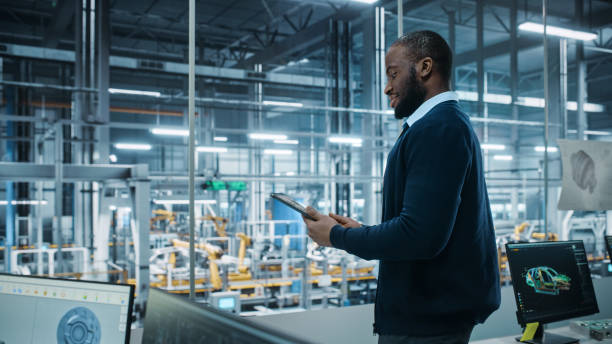 istock Car Factory Office: Portrait of Successful Black Male Chief Engineer Using Tablet Computer in Automated Robot Arm Assembly Line Manufacturing High-Tech Electric Vehicles. Side View Shot 1352825154