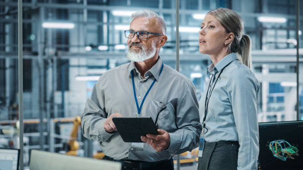istock Car Factory: Female Project Manager Talks to Male Automotive Engineer, Plan Strategy Using Tablet Computer. Automated Robot Arm Assembly Line Production Conveyor Manufacturing Electric Vehicles 1352825168