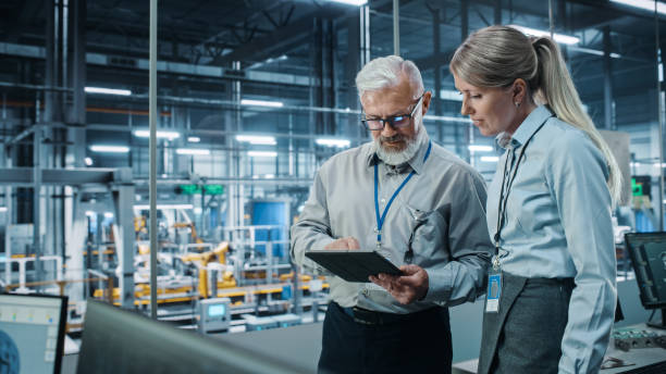 Car Factory: Female Project Manager Talks to Male Automotive Engineer, Plan Strategy Using Tablet Computer. Automated Robot Arm Assembly Line Production Conveyor Manufacturing Electric Vehicles