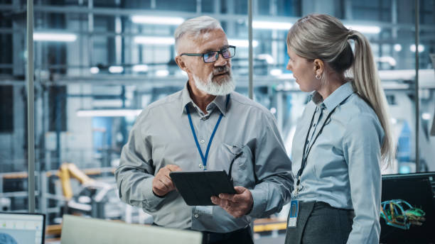 istock Car Factory: Female Project Manager Talks to Male Automotive Engineer, Plan Strategy Using Tablet Computer. Automated Robot Arm Assembly Line Production Conveyor Manufacturing Electric Vehicles 1352825172