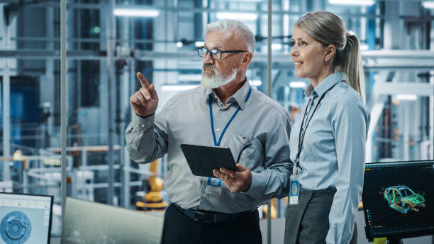 istock Car Factory: Female Project Manager Talks to Male Automotive Engineer, Plan Strategy Using Tablet Computer. Automated Robot Arm Assembly Line Production Conveyor Manufacturing Electric Vehicles 1352825174