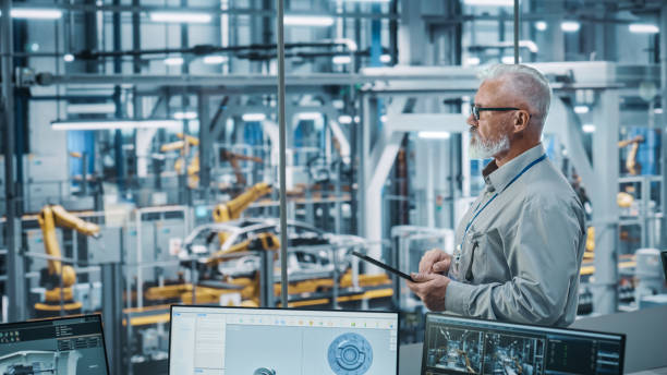 istock Car Factory Office: Portrait of Senior White Male Chief Engineer Using Tablet Computer in Automated Robot Arm Assembly Line Manufacturing High-Tech Electric Vehicles. Side View Shot 1352825196