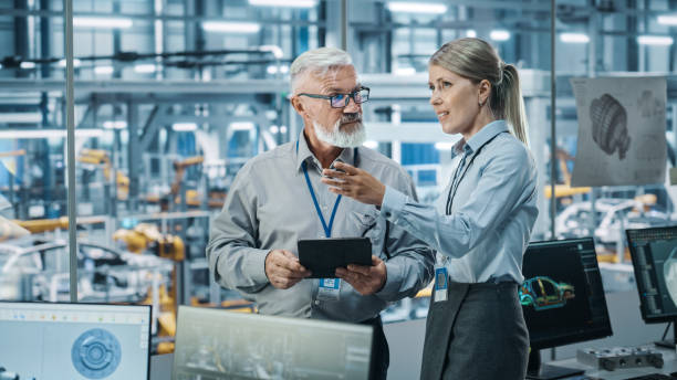istock Car Factory: Female Project Manager Talks to Male Automotive Engineer, Plan Strategy Using Tablet Computer. Automated Robot Arm Assembly Line Production Conveyor Manufacturing Electric Vehicles 1352825208