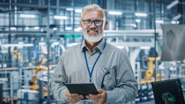istock Car Factory Office: Portrait of Senior White Male Chief Engineer Using Tablet Computer in Automated Robot Arm Assembly Line Manufacturing High-Tech Electric Vehicles. Looking at Camera and Smiling 1352825213
