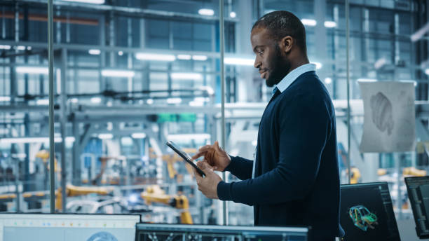 istock Car Factory Office: Portrait of Successful Black Male Chief Engineer Using Tablet Computer in Automated Robot Arm Assembly Line Manufacturing High-Tech Electric Vehicles. Medium Side View 1352825217