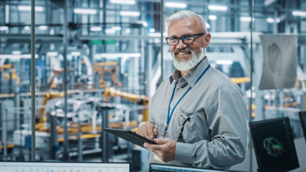 istock Car Factory Office: Portrait of Senior White Male Chief Engineer Using Tablet Computer in Automated Robot Arm Assembly Line Manufacturing High-Tech Electric Vehicles. Looking at Camera and Smiling 1352825228
