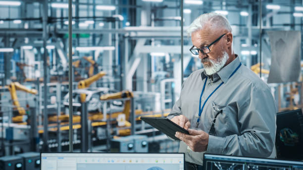 istock Car Factory Office: Portrait of Senior White Male Chief Engineer Using Tablet Computer in Automated Robot Arm Assembly Line Manufacturing High-Tech Electric Vehicles. Medium Shot 1352825231