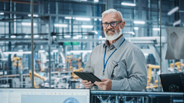 istock Car Factory Office: Portrait of Senior White Male Chief Engineer Using Tablet Computer in Automated Robot Arm Assembly Line Manufacturing High-Tech Electric Vehicles. Looking at Camera and Smiling 1352825232