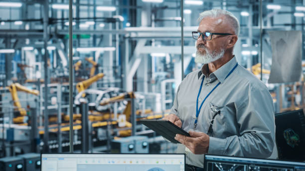 istock Car Factory Office: Portrait of Senior White Male Chief Engineer Using Tablet Computer in Automated Robot Arm Assembly Line Manufacturing High-Tech Electric Vehicles. Medium Shot 1352825261