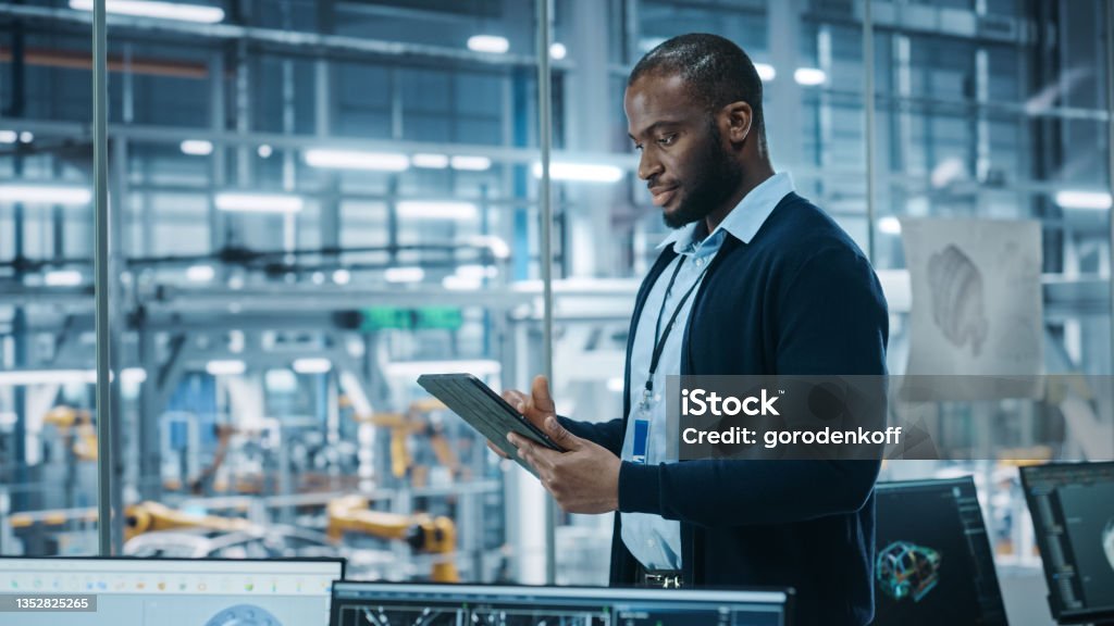 Car Factory Office: Portrait of Successful Black Male Chief Engineer Using Tablet Computer in Automated Robot Arm Assembly Line Manufacturing High-Tech Electric Vehicles. Side View Shot Industry Stock Photo