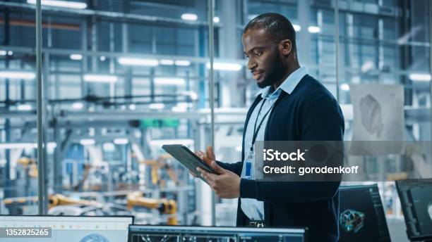 Car Factory Office Portrait Of Successful Black Male Chief Engineer Using Tablet Computer In Automated Robot Arm Assembly Line Manufacturing Hightech Electric Vehicles Side View Shot Stock Photo - Download Image Now