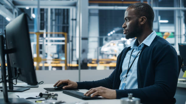istock Car Factory Office: Portrait of Confident Black Male Chief Engineer Working on Desktop Computer. Professional Technician in Automated Robot Arm Assembly Line Manufacturing High-Tech Electric Vehicles 1352825342