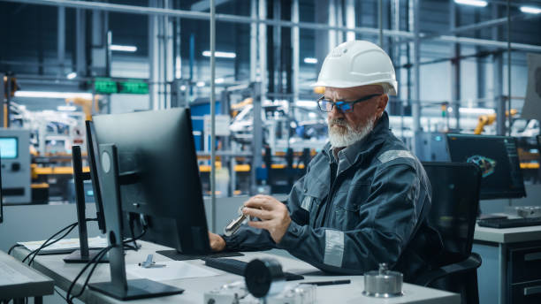 istock Car Factory Office: Portrait of Male Engineer Wearing Safety Hard Hat Working on Computer. Techniciancian Constructing Component. Automated Robot Arm Assembly Line Manufacturing Electric Vehicles 1352825367
