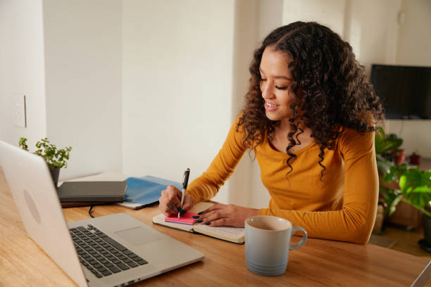 istock Multi-cultural female smiling, writing in notebook. Young professional remote working with laptop in modern apartment 1352825637