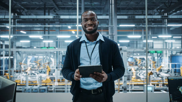 istock Car Factory Office: Portrait of Successful Black Male Chief Engineer Using Tablet Computer in Automated Robot Arm Assembly Line Manufacturing High-Tech Electric Vehicles. Medium Looking at Camera 1352825782