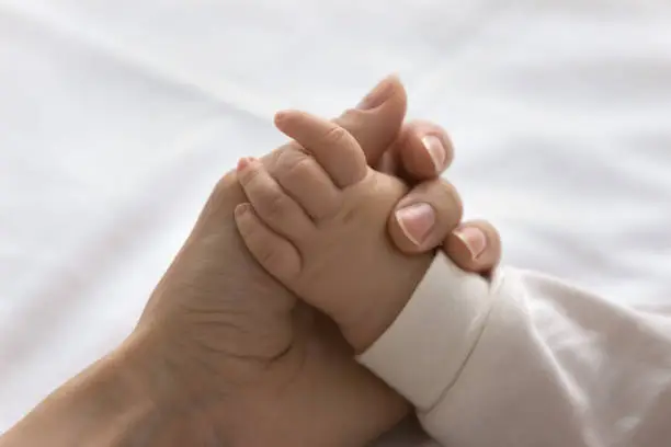 Young mom touching little palm of little baby. Mother holding cute hand of infant son or daughter in white garment. Close up, cropped shot. Motherhood support, childbirth, infancy concept