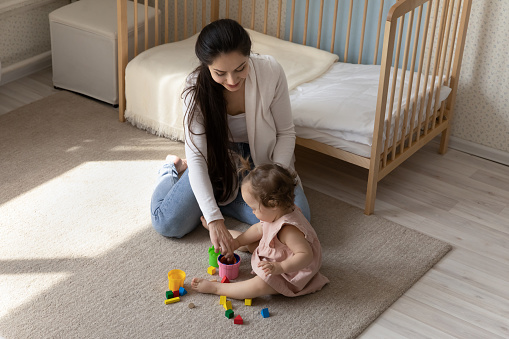 Happy loving mom and sweet toddler baby playing learning games with plastic toys on warm heating floor, caring of child development. Mother enjoying maternity leave with cute little daughter girl.