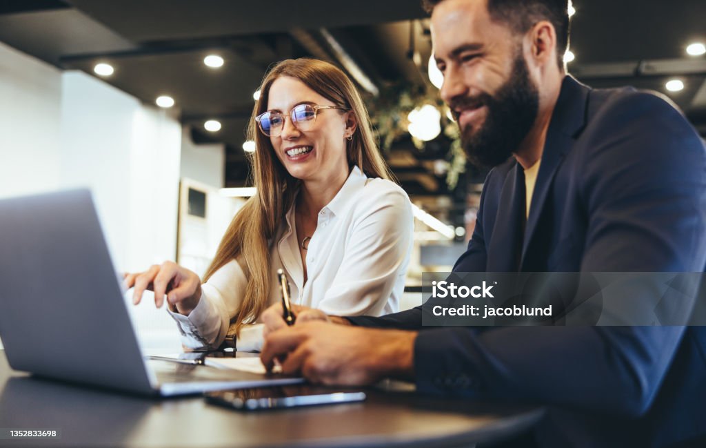 Cheerful businesspeople using a laptop in an office Cheerful businesspeople using a laptop in an office. Happy young entrepreneurs smiling while working together in a modern workspace. Two young businesspeople sitting together at a table. Office Stock Photo