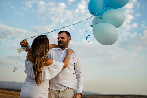 Pregnant woman and her husband revealing baby gender with blue balloons on a sunny summer day in nature