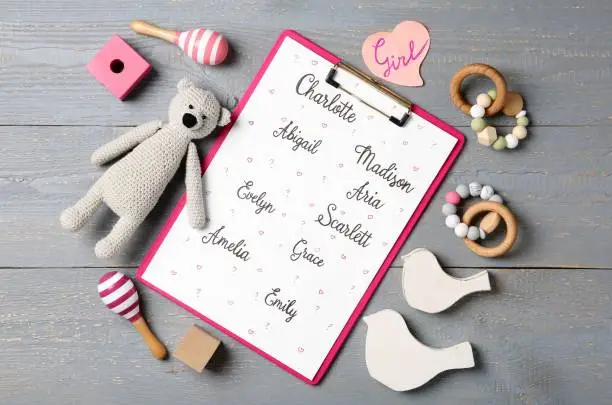 Photo of Clipboard with different baby names and toys on grey wooden table, flat lay