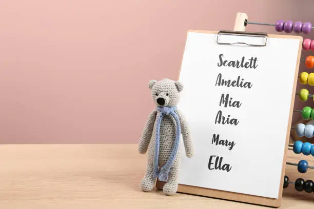 Photo of Clipboard with list of baby names and toys on wooden table. Space for text