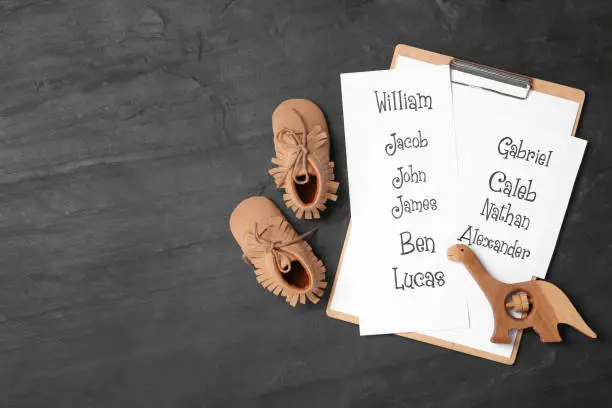 Photo of Clipboard with list of baby names, child's shoes and wooden toy on black table, flat lay. Space for text