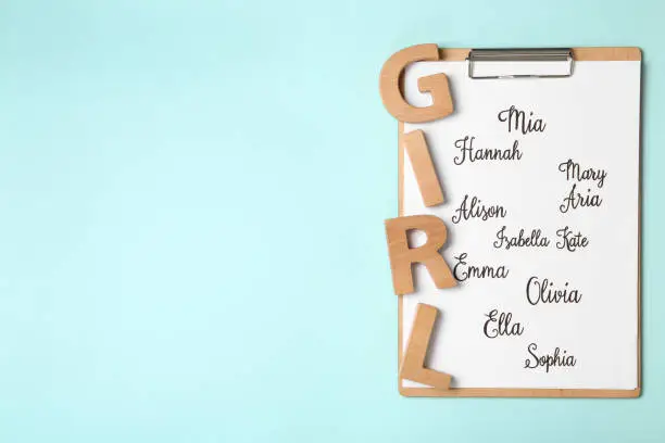 Photo of Clipboard with list of baby names and wooden letters on light blue background, top view. Space for text
