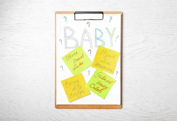 Photo of Clipboard with written different baby names on white wooden table, top view