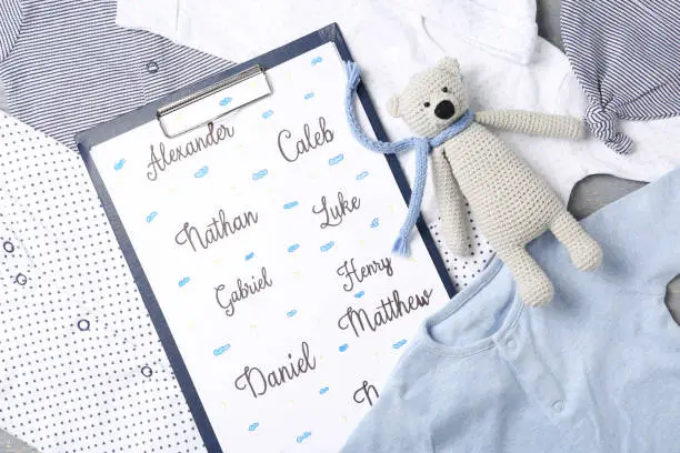 Photo of Clipboard with different baby names and toy on child's clothes, top view
