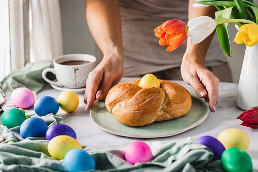 Close-up of woman serving breakfast on beautifully decorated table for Easter. Woman placing bread nest with egg on table with multicolored Easter eggs and flowers.