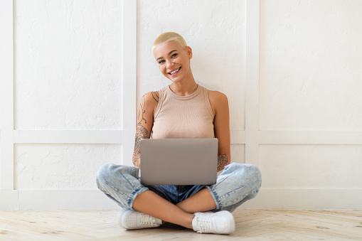 Young lady sitting on floor with laptop computer over white wall, looking at camera and smiling, copy space. Millennial woman studying or working with notebook