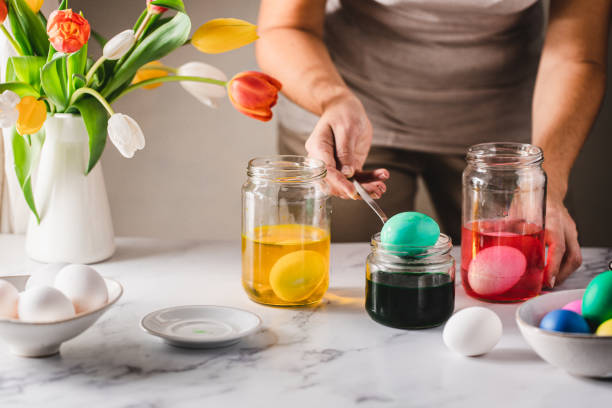 Woman dyeing Easter eggs at home Dyeing Easter eggs. Close-up of woman's hand dipping egg in bottle of dye over table in kitchen. Female coloring easter eggs with natural dye at home. dye stock pictures, royalty-free photos & images