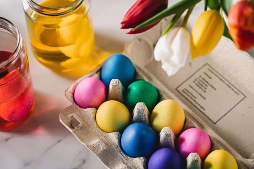 Naturally dyed Easter eggs in a carton with bottle of dye over table. Colorful Easter eggs in a carton with her cellphone. Hands of female holding mobile phone with