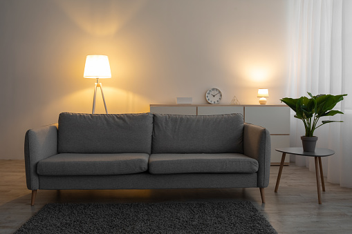 Couch, clock, plant in pot on table and glowing lamp in evening on gray wall background in living room. Modern blog about interiors, minimalistic design and retro comfortable furniture, copy space