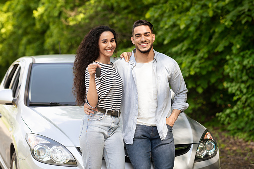 Cheerful beautiful middle-eastern couple young man and woman standing outside by nice auto, hugging and showing key from automobile, having car trip together. Car renting, leasing, sharing concept